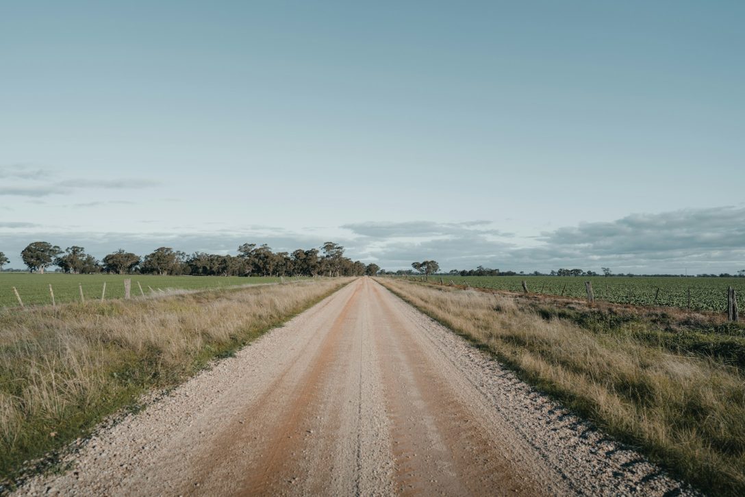 A dirt road running between two grassy fields. Photo by Arun Clarke on Unsplash